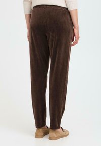 Brown corduroy pants with vertical ribbing, elastic waistband, and tapered legs. Worn with neutral slip-on shoes, viewed from the back.