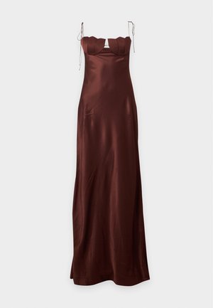 Femme grande aux longs cheveux foncés, portant une robe en satin marron sans manches avec de fines bretelles et des bonnets festonnés, debout devant un fond uni.
