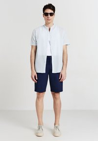 Uomo che indossa occhiali da sole neri, una camicia a maniche corte azzurro chiaro sopra una maglietta bianca, pantaloni corti blu navy e scarpe in camoscio beige su sfondo bianco.