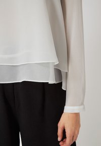 Blusa blanca, transparente, con un dobladillo en capas y mangas largas; presenta un puño abotonado. Llevada sobre pantalones de tela negra. Material suave y ligero.