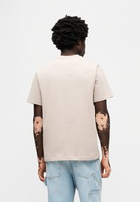 Μπεζ βαμβακερή μπλούζα με κοντά μανίκια, στρογγυλή λαιμόκοψη και άνετη εφαρμογή, φορεμένη με τζιν παντελόνι ανοιχτού γαλάζιου χρώματος. Διακριτική υφή, μινιμαλιστικός σχεδιασμός.