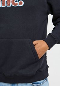 Sudadera negra con un logotipo texturizado y de color en el frontal. Presenta un bolsillo canguro y puños acanalados. Fabricada en mezcla de algodón.