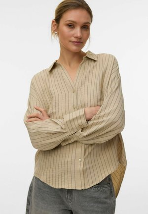 Femme aux cheveux châtain clair portant une chemise beige à manches longues à rayures verticales et un jean gris, debout les bras croisés devant un fond uni.
