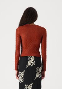 Maglione lungo a maniche con tessuto ruggine e texture, dal taglio aderente, con scollatura rotonda e dettagli a costine, abbinato a una gonna nera a fantasia.