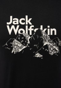 Schwarzes T-Shirt mit weißen Schriftzug "Jack Wolfskin" und einem Berggrafik. Das Design enthält komplexe Liniendetails und Texturen.