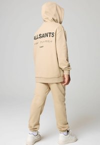 Beżowa bluza z kapturem i dopasowane joggersy, z dużym czarnym logo "ALLSAINTS" na plecach. Gładki materiał z prążkowanymi mankietami i dołem.