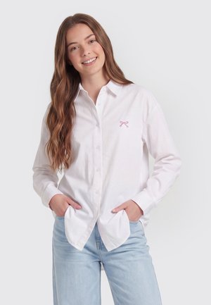Junge Frau mit langen braunen Haaren lächelt, trägt ein weißes Hemd mit Knöpfen und eine kleine rosa Schleife auf der Brust sowie hellblaue Jeans.