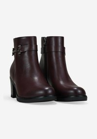 Un par de botas de cuero marrón oscuro hasta el tobillo con tacones cuadrados, cremalleras laterales y correas decorativas con hebillas plateadas sobre un fondo blanco.