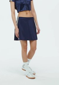 Mornarska atletska suknja od glatkog materijala, s bijelim bočnim trakama. Uparena s bijelim tenisicama i čarapama do koljena.