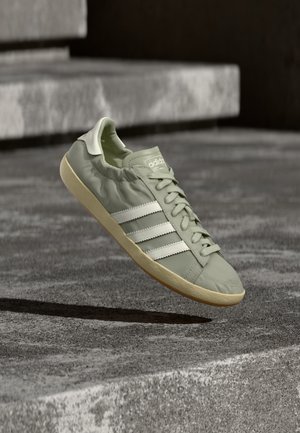 Baskets Adidas gris clair avec des bandes blanches et une semelle beige flottant au-dessus des marches en béton.