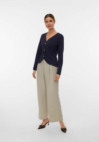 Mornarski jopič na gumbe z V-izrezom, dolgimi rokavi in gladkim materialom. Ujemajo se s svetlo sivo širokima hlačami in črnimi koničastimi petami.