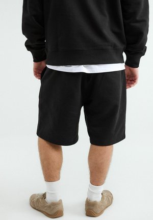 Osoba ubrana w czarne spodenki, czarną bluzę, białe skarpetki i beżowe tenisówki, stojąca tyłem na jednolitym białym tle.