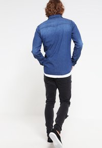 Koszula denimowa w ciemnoniebieskim kolorze, z klasycznym kołnierzykiem, długimi rękawami i zapinanymi na guziki mankietami, zestawiona z czarnymi dżinsami i białymi trampkami.