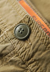 Zbliżenie na materiał khaki z okrągłym zielonym guzikiem z napisem "SUPERDRY" i "PARACHUTE", szczegółami szwów oraz gładką teksturą.