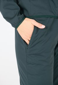 Donkergroen buitenwear heeft een soepele textuur, een ritszak aan de zijkant en elastische manchetten voor een aangepaste pasvorm en comfort.