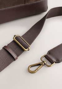 Brown fabric and leather adjustable bag strap with brass swivel clasp and slider on white surface.