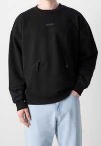 Felpa nera Hugo con tasche con cerniera indossata da persona con jeans azzurri, una mano in tasca e l'altra rilassata di fianco.