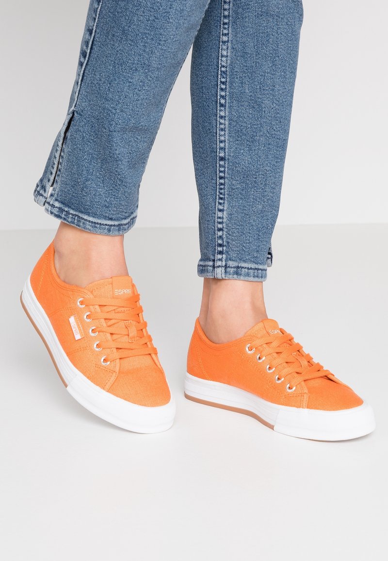 Personne portant un jean bleu et des baskets en toile orange vif avec des semelles blanches, debout sur un sol gris clair.