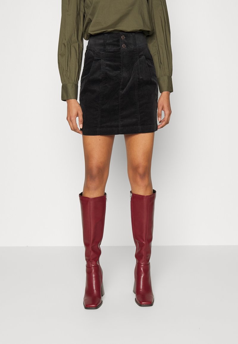 Femme portant une mini-jupe noire en velours côtelé taille haute avec poches devant, un chemisier vert olive et des bottes en cuir bordeaux montantes jusqu’aux genoux.