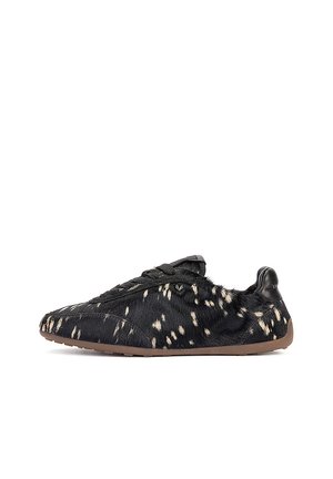 Zapatilla negra de corte bajo con patrón de piel manchada blanca y suela marrón mostrada de lado sobre fondo blanco.