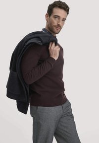 Burgunderfarbener Strickpullover mit ribbeltem Saum; graues Karohemd darunter; hält ein dunkelblaues Sakko über der Schulter; heller Hintergrund.