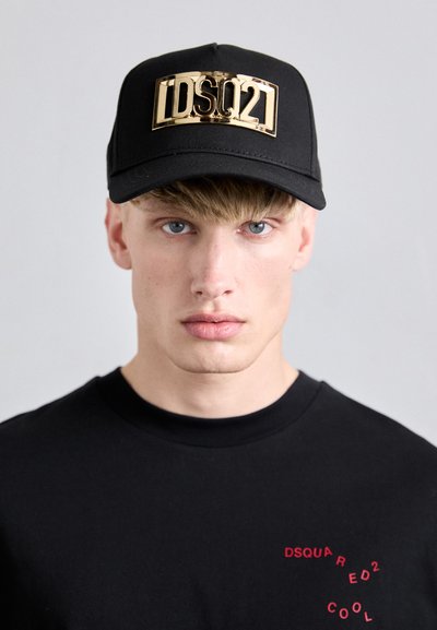 Schwarzne Baseballmütze mit einer goldenen Logoplatte, kombiniert mit einem schwarzen T-Shirt mit rotem Text. Die Mütze hat einen gebogenen Schirm und einen verstellbaren Riemen.