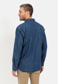 Denimskjorte i mørkeblå med lange ærmer, knaplukning ved manchetterne og spids krave. Har en løkke i den bageste halsudskæring. Casual pasform.