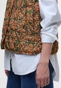 Quilted green vest adorned with intricate orange floral patterns. Features a side pocket and logo detailing. Paired with a white shirt.