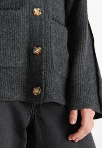 Tmavě šedá texturovaná bunda se čtyřmi velkými kulatými knoflíky a přední kapsou. Tkanina má pruhovaný vzhled, který vytváří hloubku.