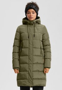 Žena u maslinasto zelenom zimskoj kaputu s kapuljačom i crnom krznenom trakom za glavu, stoji očevidno nasuprot jednostavne svijetle pozadine.