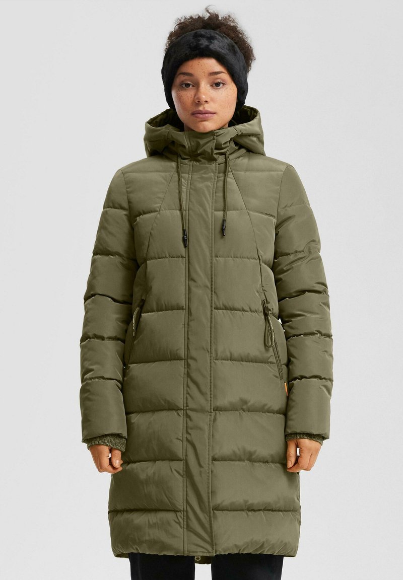 Žena u maslinasto zelenom zimskoj kaputu s kapuljačom i crnom krznenom trakom za glavu, stoji očevidno nasuprot jednostavne svijetle pozadine.