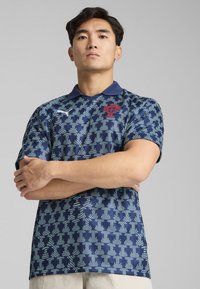 Polo shirt bleu marine avec un motif géométrique texturé en bleu et blanc, doté d'un col et d'un emblème rouge sur la poitrine.