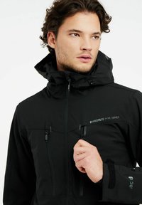 Schwarze wasserdichte Jacke mit hohem Kragen, Reißverschlusstaschen, verstellbarem Kapuze und taktilen Akzenten; mit Markenlogo auf der Vorderseite.