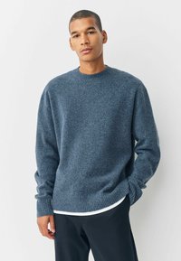 Jonge man in een blauwe gebreide trui over een wit shirt en zwarte broek, staand met een hand in zijn zak tegen een egale achtergrond.