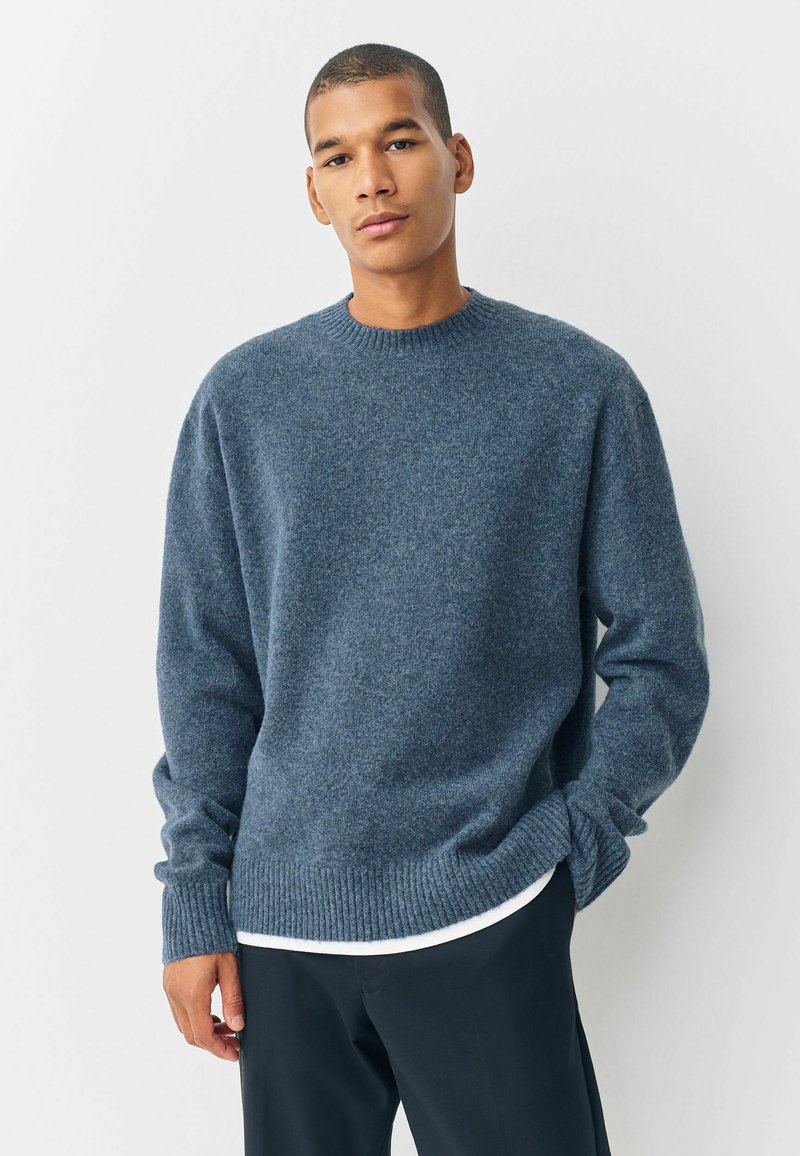 Jonge man in een blauwe gebreide trui over een wit shirt en zwarte broek, staand met een hand in zijn zak tegen een egale achtergrond.