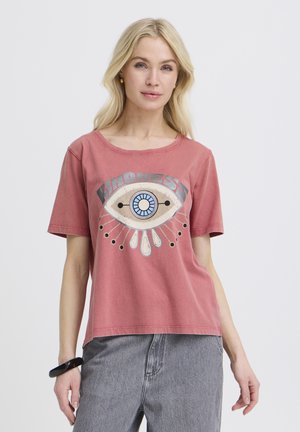 Blond kvinde iført en falmet rød T-shirt med en stor øje-grafik og ordet 'KINDNESS', kombineret med grå jeans og et sort armbånd.