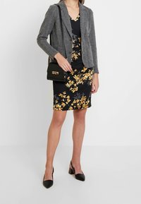 Šedý strukturovaný blazer přes černé květinové šaty s žlutými akcenty, doplněný o černé špičaté podpatky a malou černou kabelku.