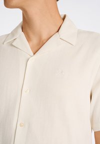 Camisa de botones de manga corta en tejido color crema claro, con cuello puntiagudo, sutil textura y logo bordado en el pecho.
