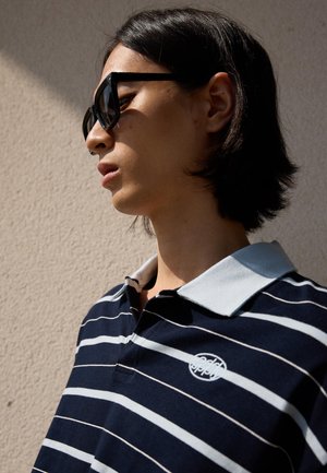 Young person with medium-length hair wearing black sunglasses and a navy striped polo shirt, standing against a beige wall.