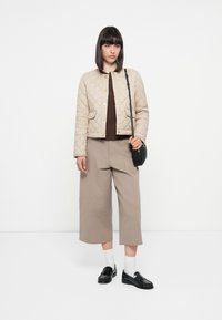 Quilted beige jacket with snap buttons, two front pockets, worn over a brown sweater and beige wide-leg pants, paired with black loafers and white socks.