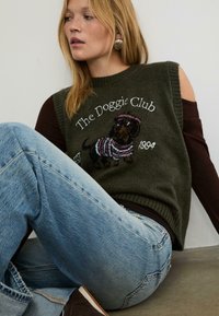 Grön stickad ärmlös tröja med en design av en tax i randig klädsel, med texten "The Doggie Club EST 1994." Matchad med blå jeans.