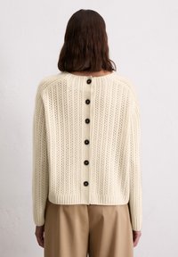 Cremfarbener Strickpullover mit vertikaler Rippung, auffälligen schwarzen Knöpfen auf der Rückseite und einer lockeren Passform. Weiche Textur mit offenem Strickmuster.