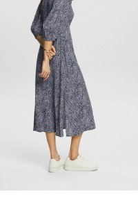 Modrá a bílá vzorovaná midi šaty s tříčtvrtečními rukávy, s vypasovaným pasem. Nosí se s bílými teniskami pro kontrast.