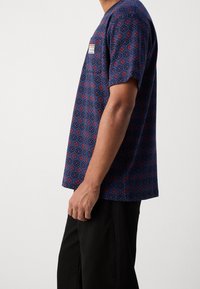 Laivastonsininen lyhythihainen paita, jossa on punainen ja sininen geometrinen kuvio. Siinä on rintatasku ja kevyt kangas.