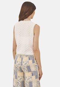 Top sin mangas de encaje blanco con bordes festoneados, combinado con pantalones anchos de patrones en tonos amarillos y azules que presentan diseños florales y geométricos.