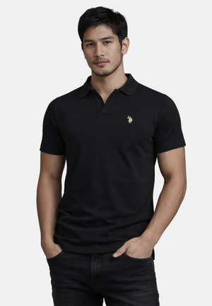 Mann trägt ein schwarzes kurzärmeliges Poloshirt mit kleinem goldenem Logo auf der Brust und schwarze Jeans, Hände in den Taschen, neutraler Hintergrund.