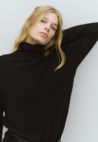 Crna dolčevita od mekog pletenog materijala, s opuštenim krojem, dugim rukavima i preklopljenim dizajnom ovratnika.