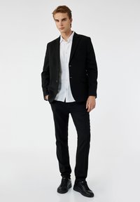 Schwarze Blazer mit einer einzigen Tasche und zwei Knöpfen, kombiniert mit einem weißen Hemd und schwarzen Hosen. Schwarze Sneakers vervollständigen das Outfit.