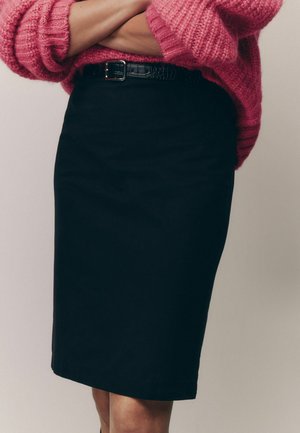 Femme portant une jupe crayon noire avec une ceinture noire et un gros pull rose tricoté, bras croisés sur la poitrine.