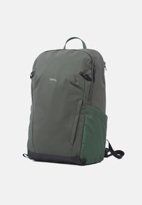 Dunkelgrüner Stoff-Rucksack mit glatter Textur, Reißverschlusstaschen, robusten Trägern und einem kontrastierenden schwarzen Boden. Logo auf der Vorderseite angebracht.